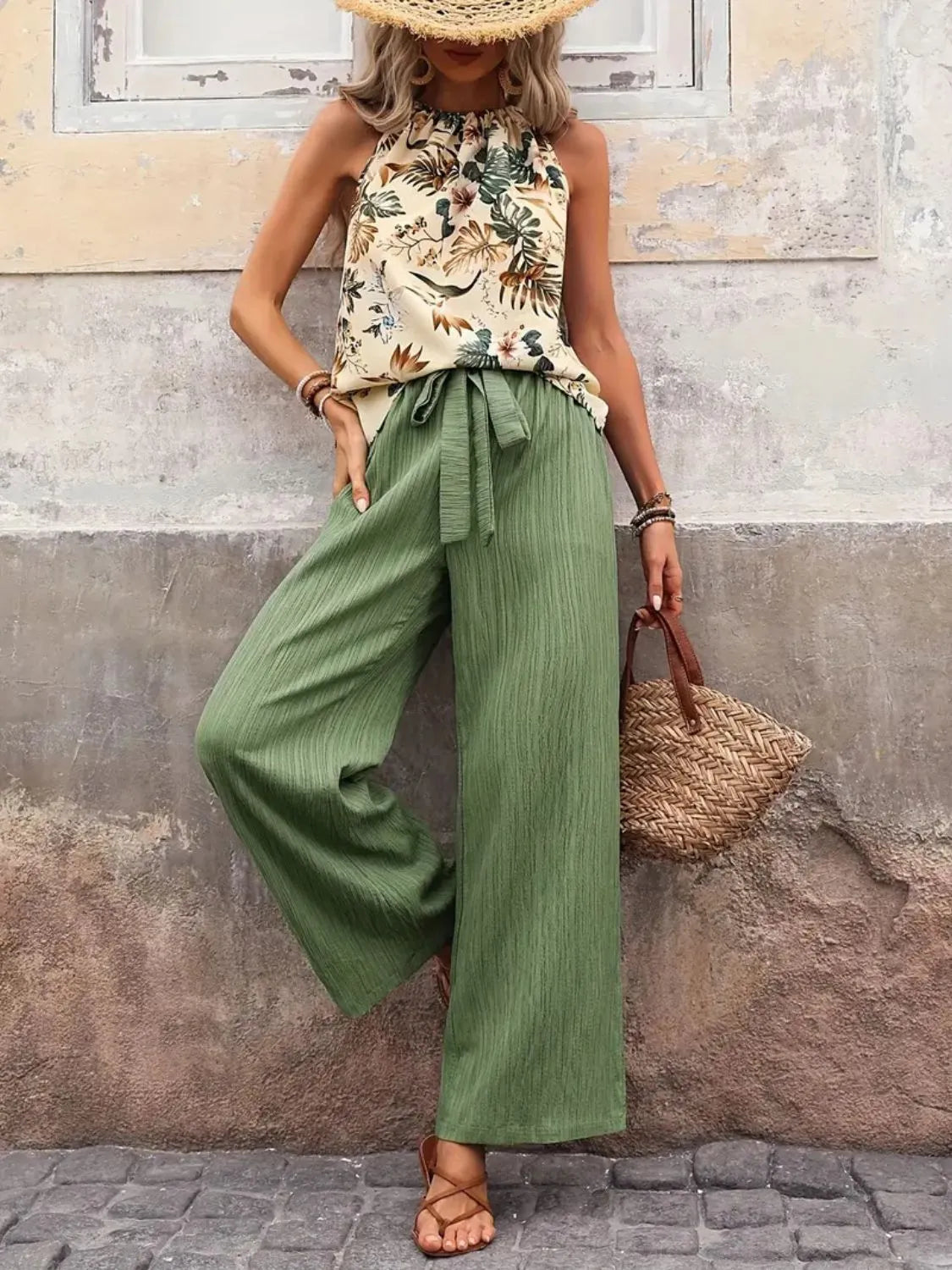 Woman wearing a floral top and green wide-leg pants against a textured wall.