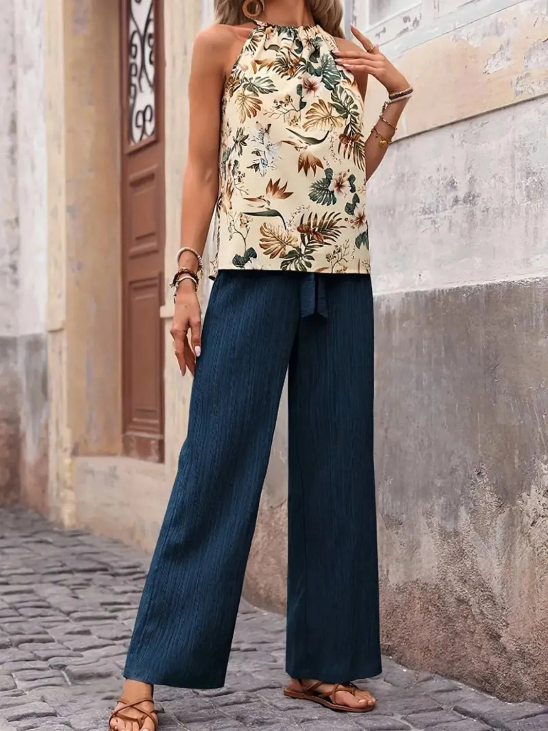 Woman wearing a floral top and blue pants standing against a textured wall.