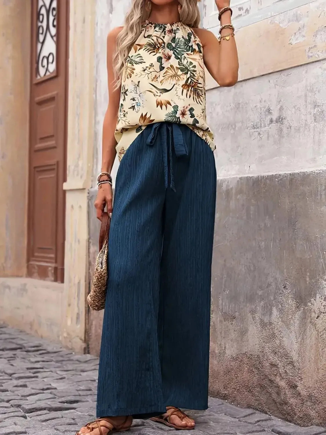 Woman wearing a floral top and blue wide-leg pants standing against a stone wall.