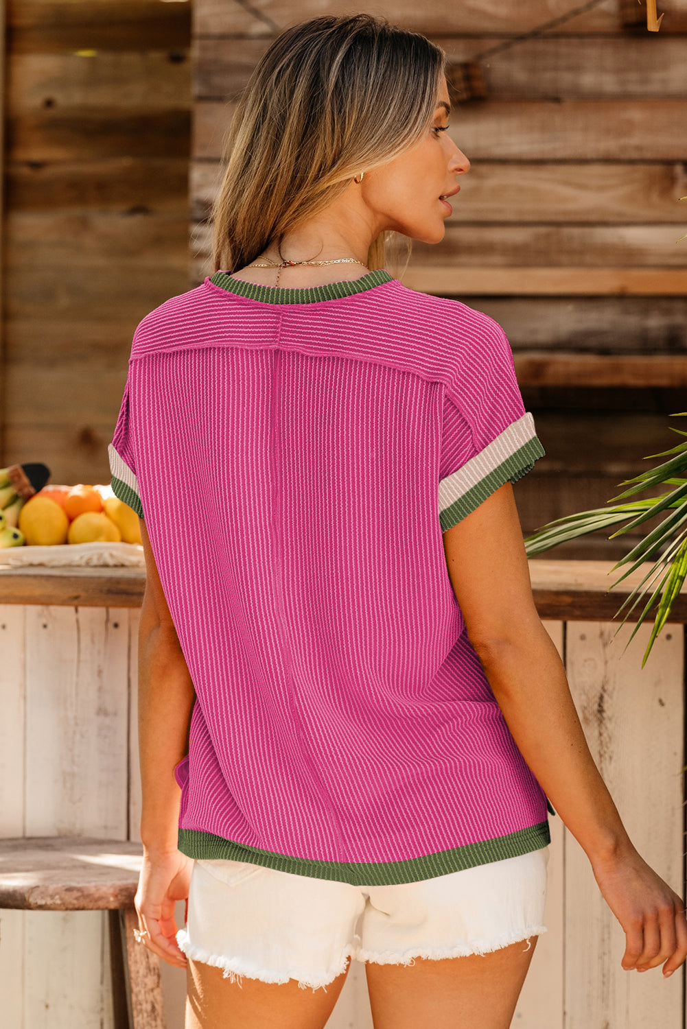 Woman wearing a pink ribbed sweater with green and white trim, standing in front of a wooden wall.
