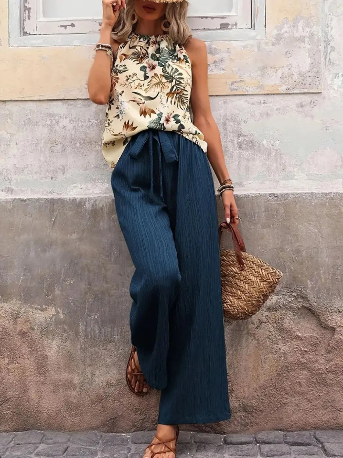 Woman wearing a floral top and blue pants with a straw bag against a textured wall.