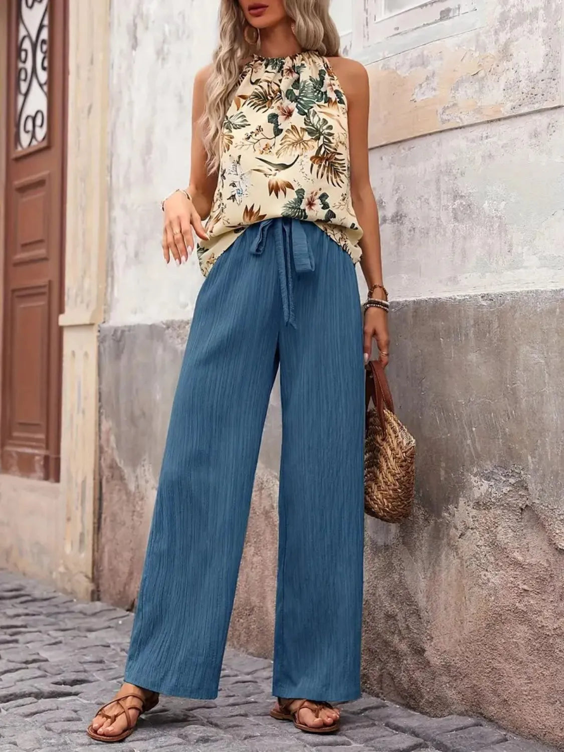 Woman wearing a floral top and blue wide-leg pants standing against a stone wall.