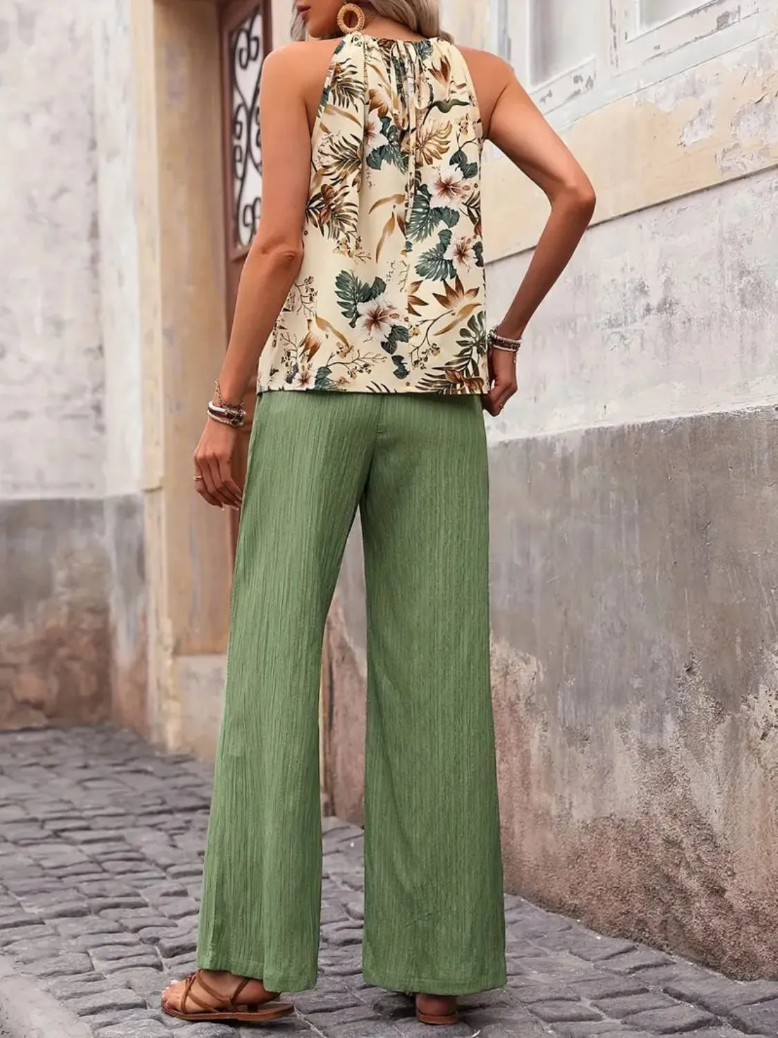 Person wearing a floral sleeveless top and green pants standing against a textured wall.