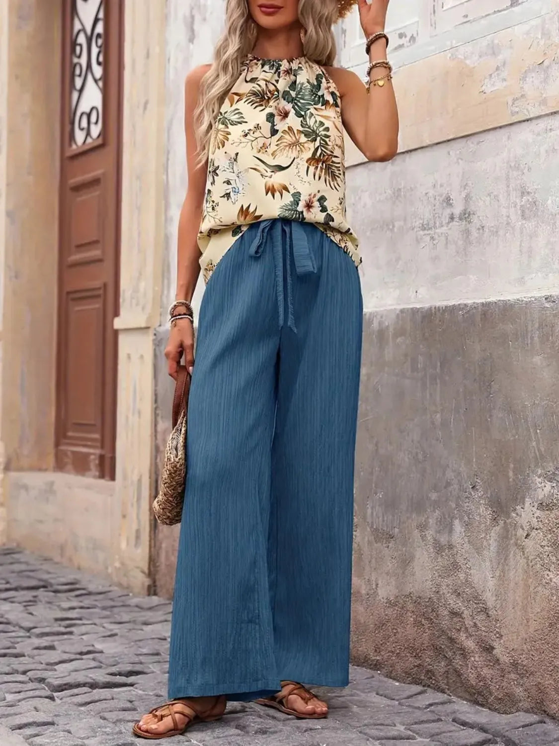 Woman wearing a floral top and blue wide-leg pants standing against a stone wall.