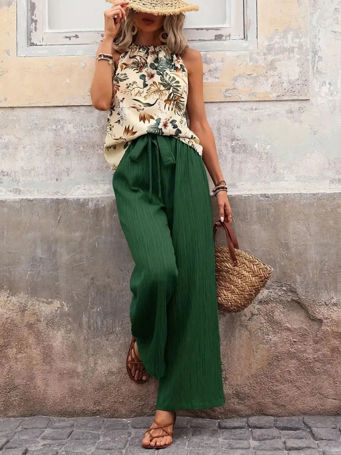Woman wearing a floral top and green pants against a textured wall.