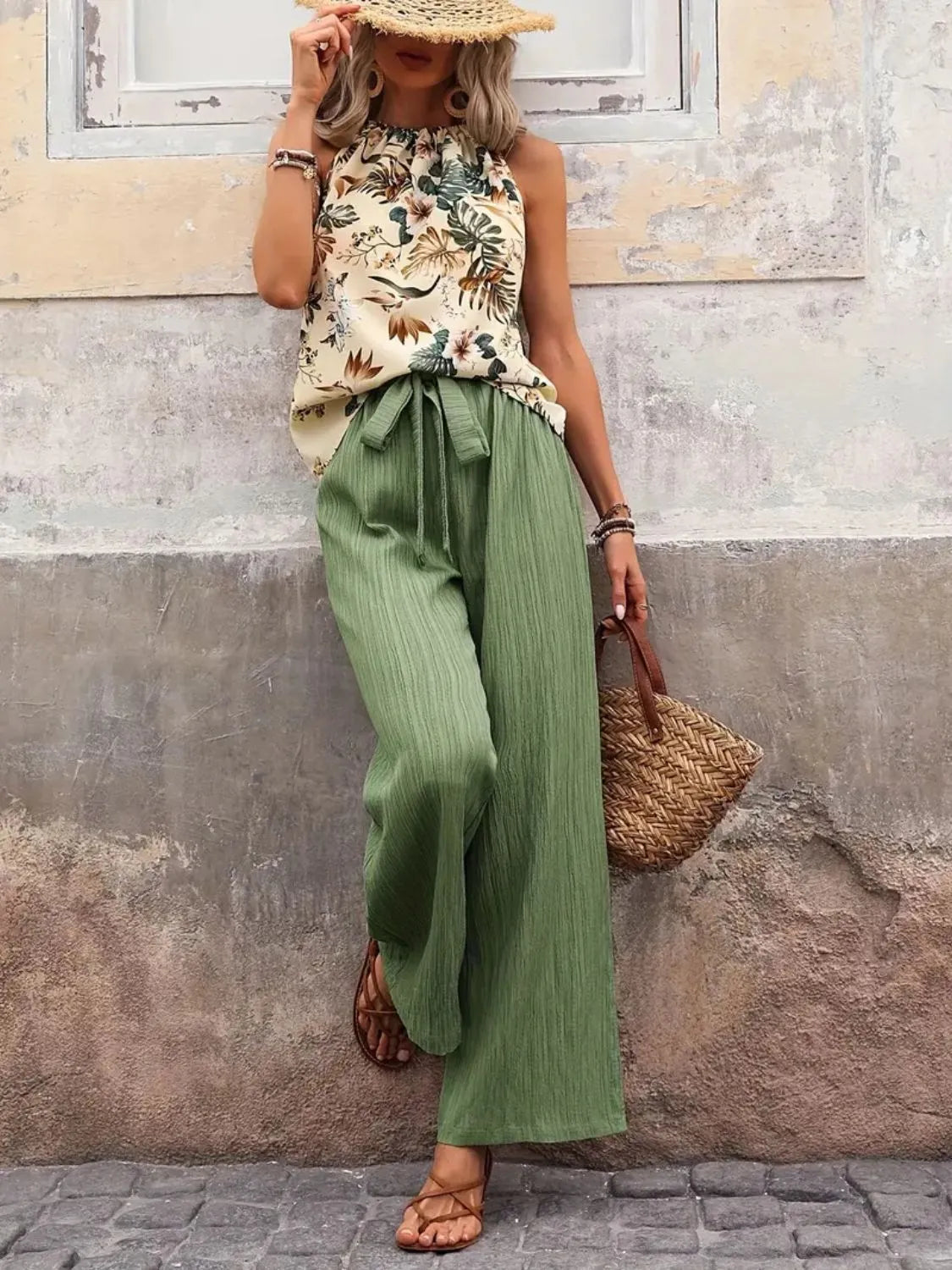 Woman wearing a floral top and green wide-leg pants against a textured wall.