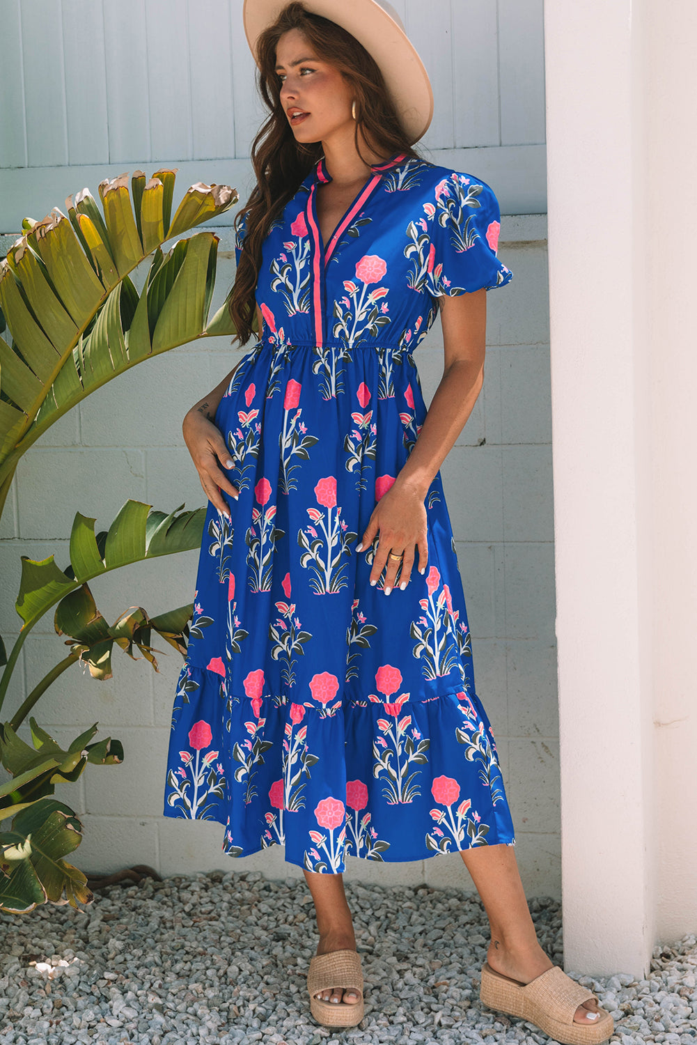 Woman wearing a blue floral dress standing outdoors with greenery and a white column in the background.