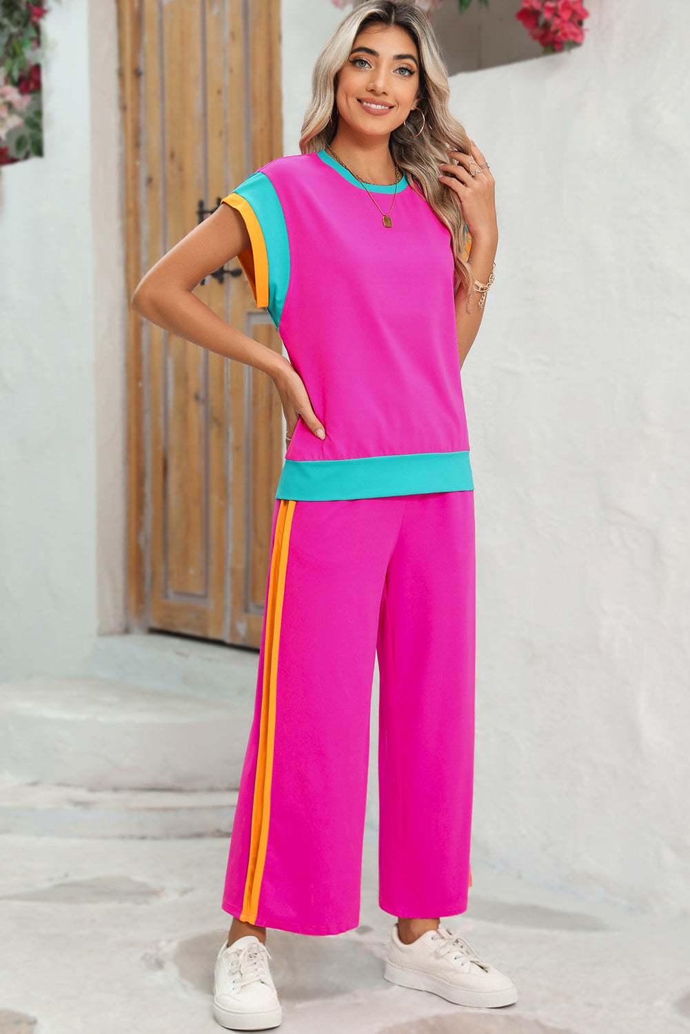 Woman wearing a bright pink outfit with colorful sleeves and pants, standing against a white wall.