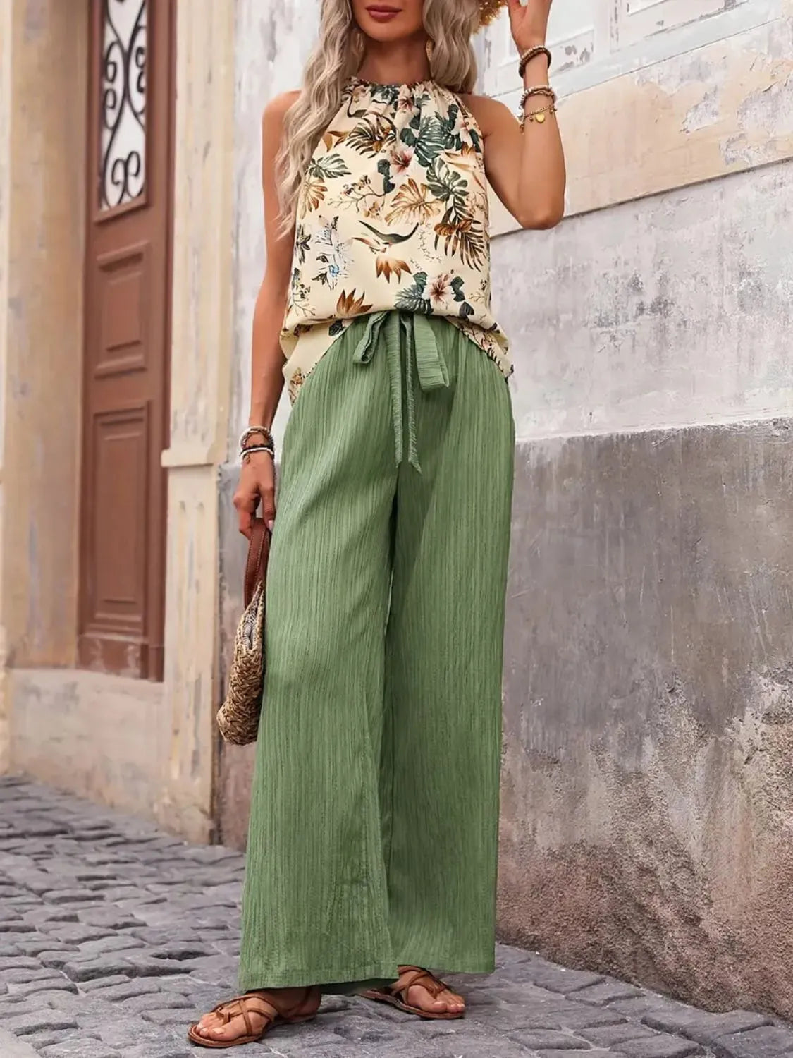 Woman wearing a floral top and green wide-leg pants standing against a textured wall.