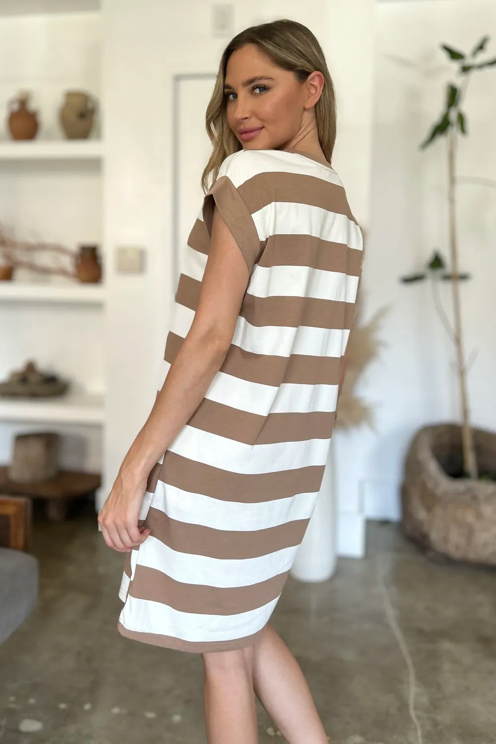 Woman wearing a brown and white striped dress in a room with a shelf and plant.