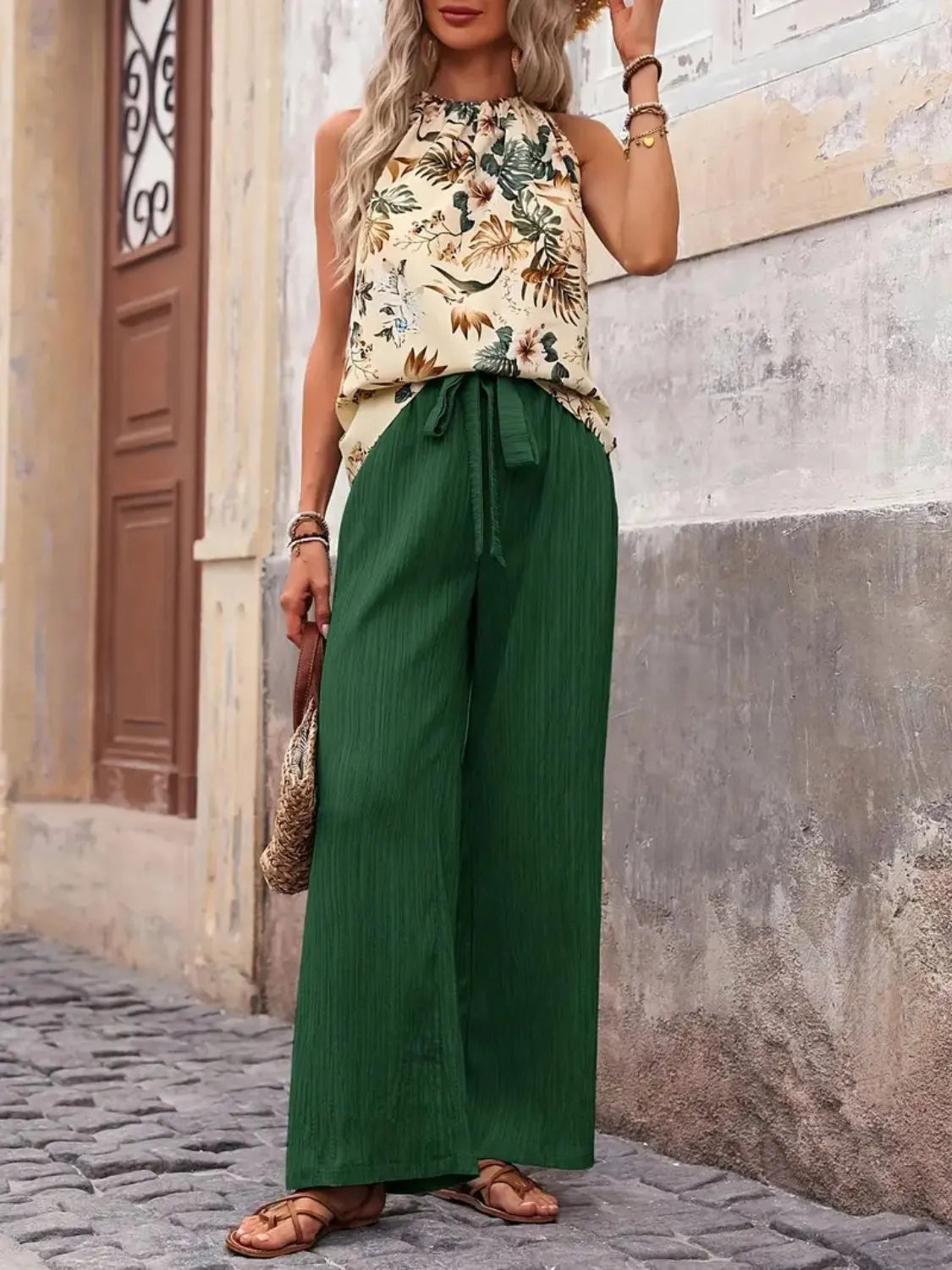 Woman wearing a floral top and green wide-leg pants standing against a textured wall.
