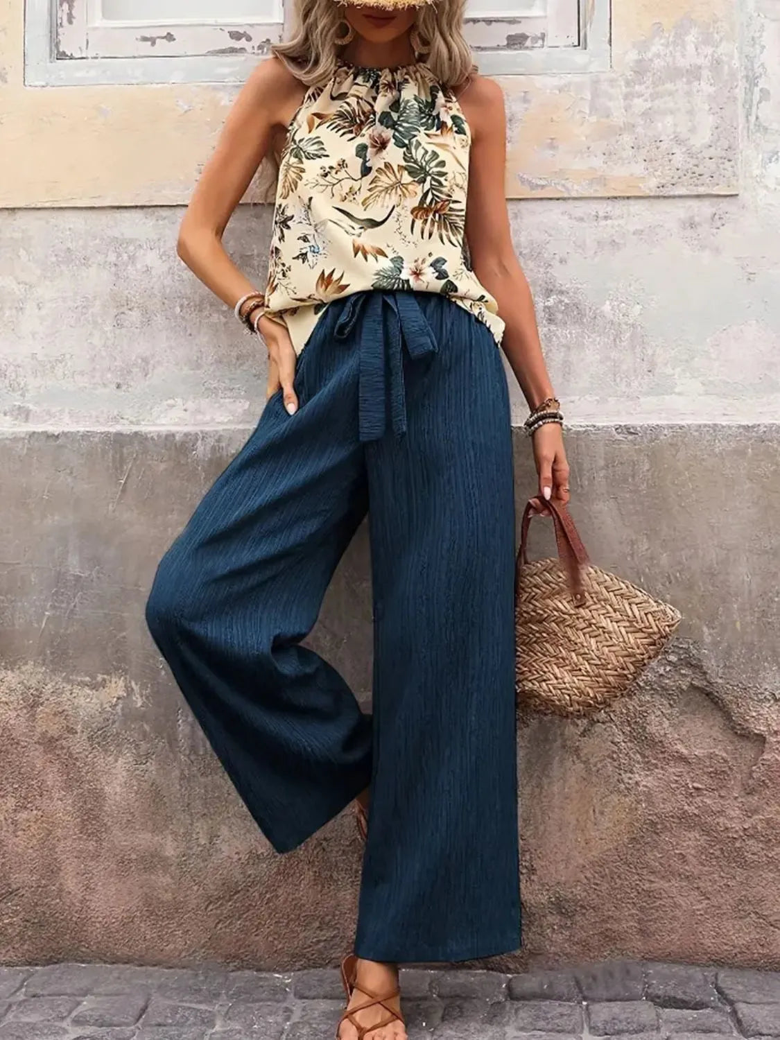 Woman wearing a floral top and wide-leg pants, holding a straw bag against a stone wall.