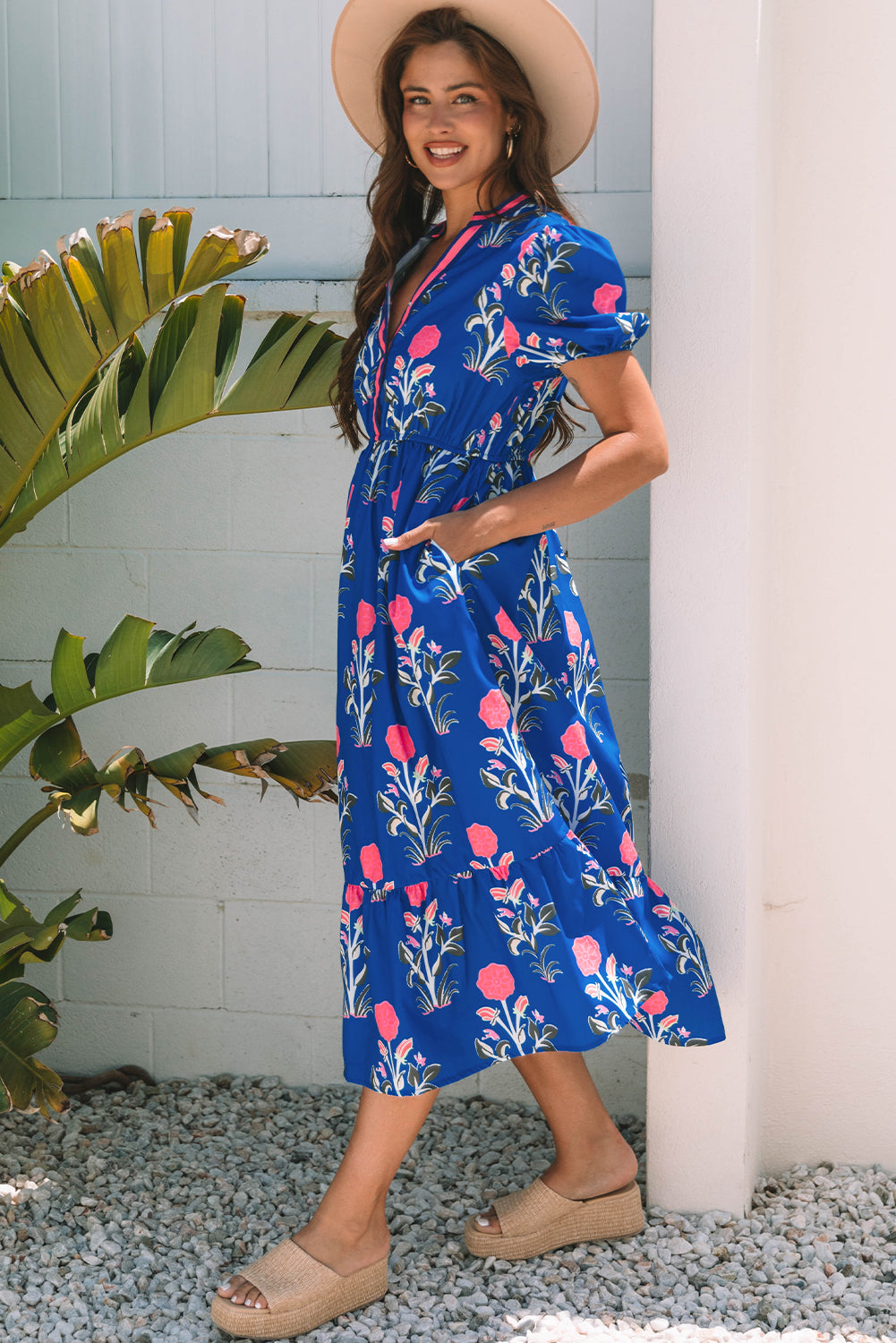 Woman wearing a blue floral dress standing outdoors with plants and a white column in the background.