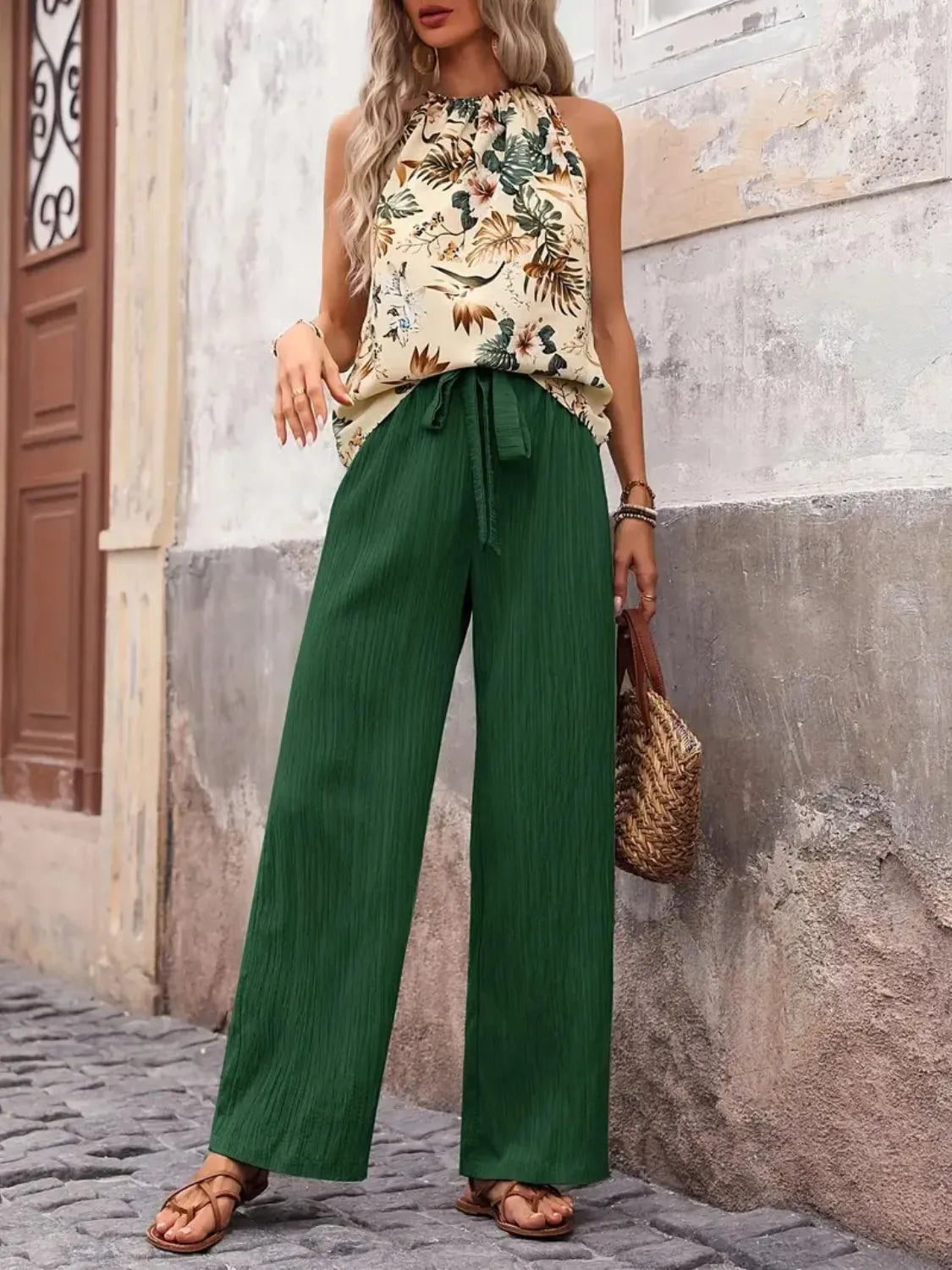 Woman wearing a floral top and green pants standing against a stone wall.