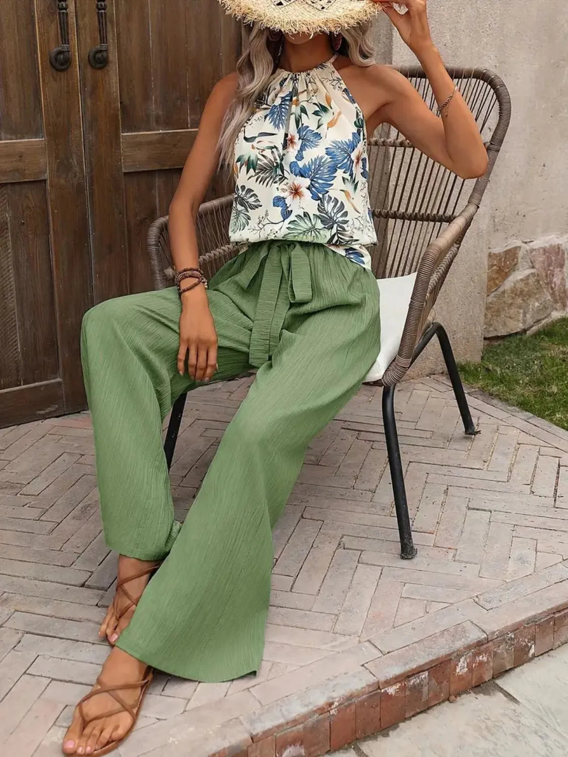 Woman sitting on a chair wearing a floral top and green pants, with a straw hat.