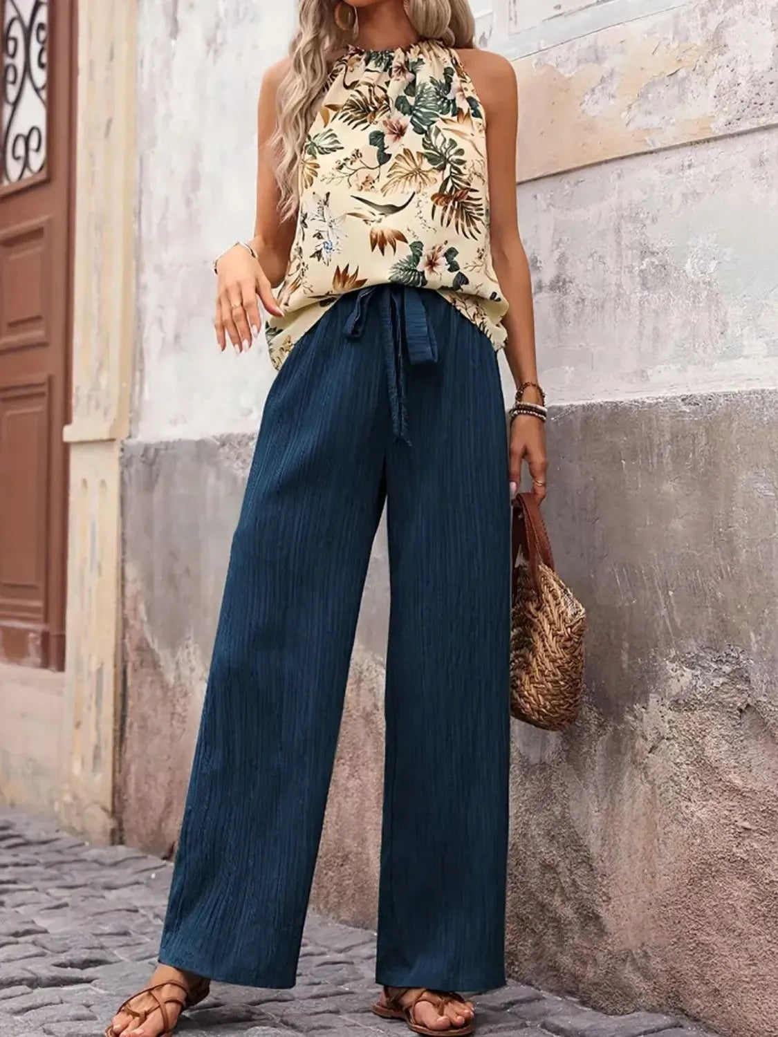 Woman wearing a floral top and blue pants standing against a textured wall.