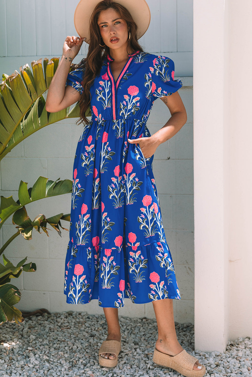 Woman wearing a blue floral dress standing outdoors with a plant and white wall in the background