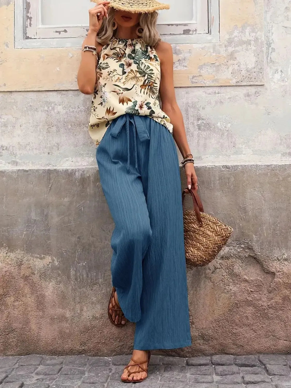 Woman wearing a floral top and blue wide-leg pants against a textured wall.