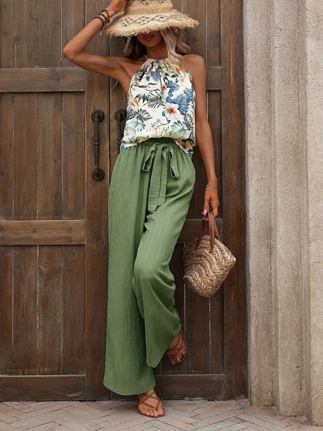 Woman in floral top and green pants standing against a wooden door.